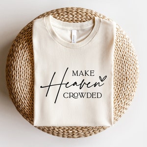 Puede incluir: Una camiseta de color crema con el texto "MAKE Heaven CROWDED" en letras negras con un peque&ntilde;o s&iacute;mbolo de coraz&oacute;n.