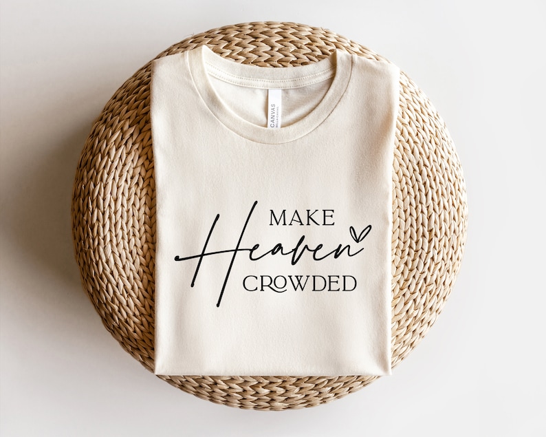 Puede incluir: Una camiseta de color crema con el texto "MAKE Heaven CROWDED" en letras negras con un peque&ntilde;o s&iacute;mbolo de coraz&oacute;n.