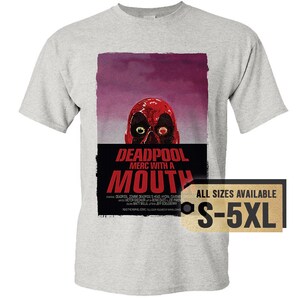 May include: A light gray t-shirt featuring a Deadpool graphic. The graphic includes a red Deadpool mask with the text "DEADPOOL MERC WITH A MOUTH". The shirt also has a tag that says "ALL SIZES AVAILABLE S-5XL".