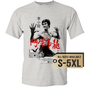 May include: A gray t-shirt with a black and white image of Bruce Lee in a fighting pose. The text "Enter the Dragon" is written in red Chinese characters on the shirt. The text "All Sizes Available S-5XL" is on a gold tag.