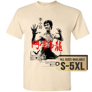 May include: A beige t-shirt with a black and white image of Bruce Lee in a fighting pose. The image is from the movie "Enter the Dragon" and includes the Chinese characters for "Enter the Dragon". The text "All Sizes Available S-5XL" is printed on a brown tag.