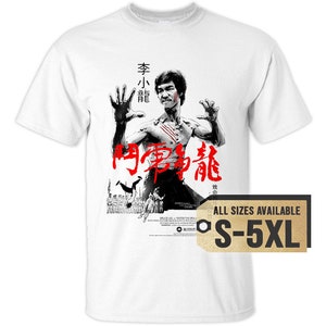 May include: White t-shirt with a black and white image of Bruce Lee in a fighting pose. The text "Enter the Dragon" is written in red Chinese characters on the shirt. The text "All Sizes Available S-5XL" is on a gold tag.