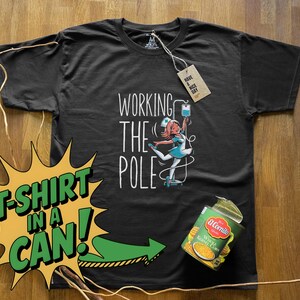 Op de afbeelding: Zwart t-shirt met de tekst "WORKING THE POLE" en een illustratie van een verpleegster. Een label leest "HAVE A NICE DAY". Een blik maïs staat naast het shirt. Het shirt is gelabeld "T-SHIRT IN A CAN!"