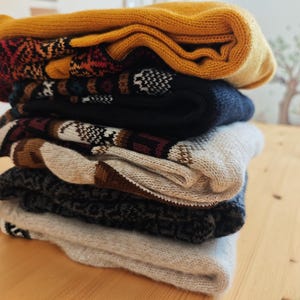 May include: A stack of folded knitwear, including socks and a hat, in various colors. The top item is mustard yellow, followed by patterned socks in red, black, and white. The stack rests on a light wooden surface.