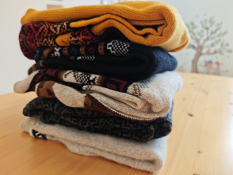 May include: A stack of folded knitwear, including socks and a hat, in various colors. The top item is mustard yellow, followed by patterned socks in red, black, and white. The stack rests on a light wooden surface.