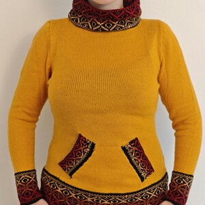 May include: Mustard yellow sweater with a patterned collar, cuffs, and pocket details. The pattern incorporates black, red, and gold geometric designs. The sweater has a turtleneck and long sleeves.