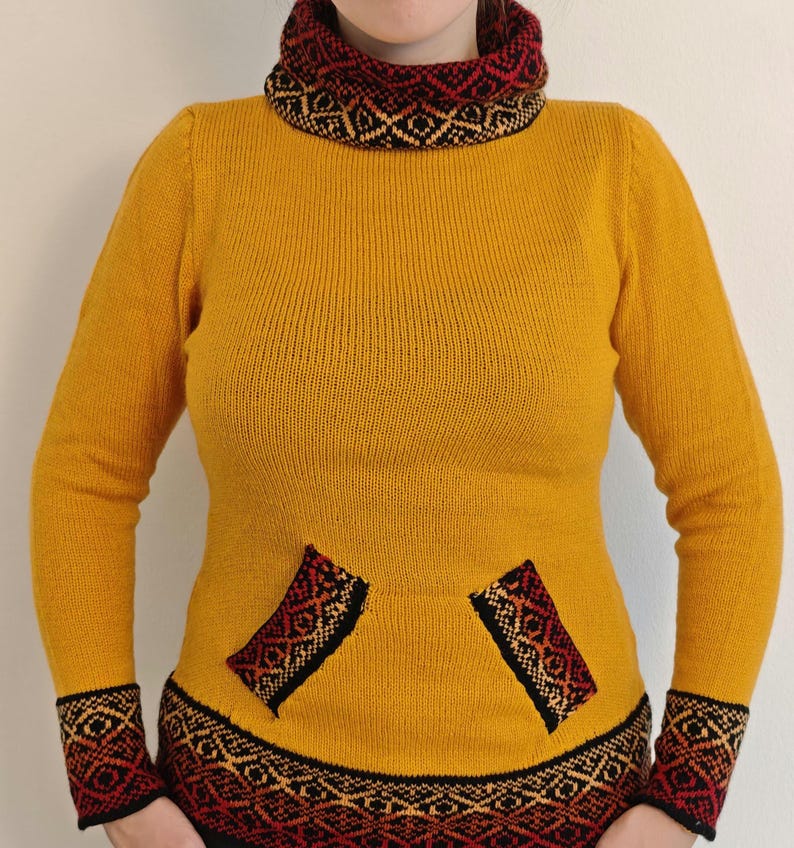 May include: Mustard yellow sweater with a patterned collar, cuffs, and pocket details. The pattern incorporates black, red, and gold geometric designs. The sweater has a turtleneck and long sleeves.
