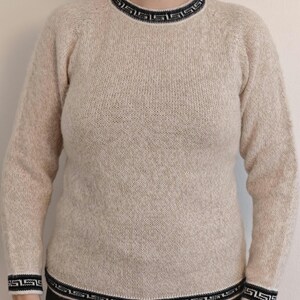 May include: A beige sweater with a crew neck and long sleeves. The neckline, cuffs, and hem feature a black and white geometric pattern. The sweater is made of a textured knit fabric.