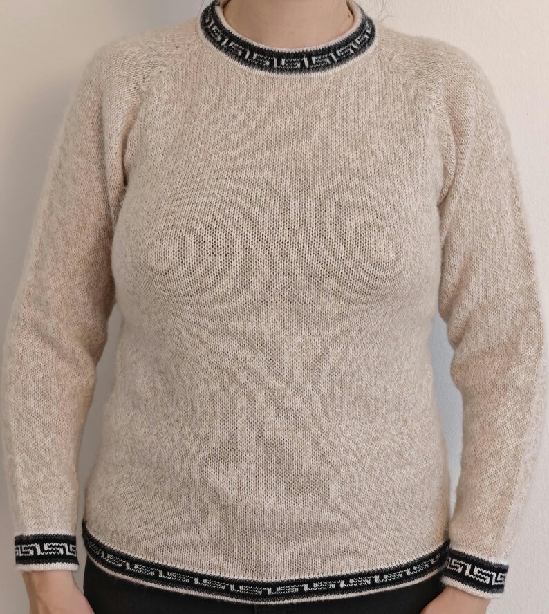 May include: A beige sweater with a crew neck and long sleeves. The neckline, cuffs, and hem feature a black and white geometric pattern. The sweater is made of a textured knit fabric.