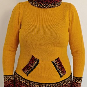 May include: Mustard yellow sweater with a patterned collar, cuffs, and pocket accents. The sweater features a high neck and long sleeves. The cuffs and collar have a black, red, and tan geometric design. The pockets also have a similar design.