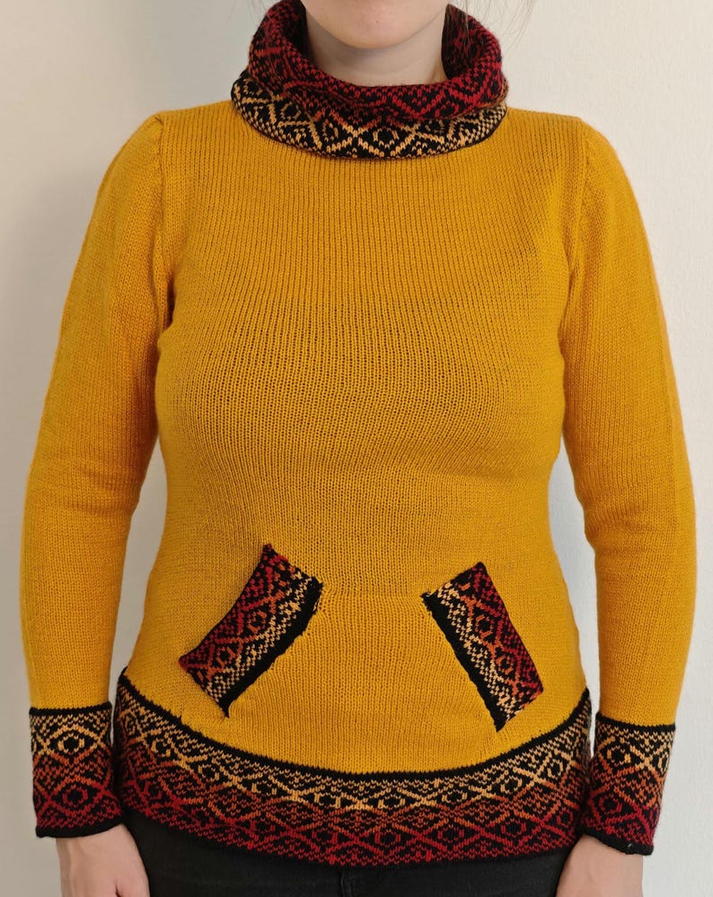 May include: Mustard yellow sweater with a patterned collar, cuffs, and pocket accents. The sweater features a high neck and long sleeves. The cuffs and collar have a black, red, and tan geometric design. The pockets also have a similar design.