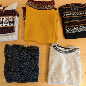 May include: Five folded sweaters with various patterns and colors are displayed on a wooden surface. The sweaters include designs with llamas, geometric shapes, and traditional knit patterns. Colors include beige, yellow, black, and gray.