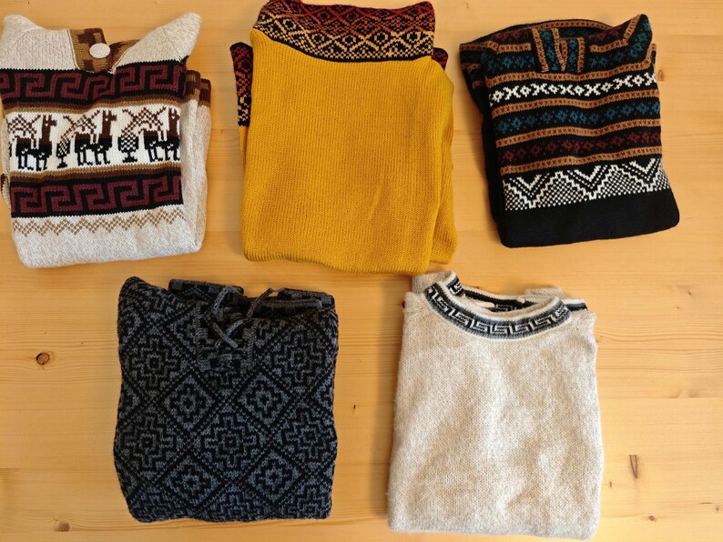 May include: Five folded sweaters with various patterns and colors are displayed on a wooden surface. The sweaters include designs with llamas, geometric shapes, and traditional knit patterns. Colors include beige, yellow, black, and gray.