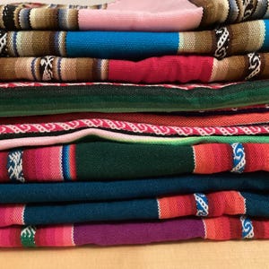 Può includere: Una pila di coperte tessute colorate. Le coperte presentano una varietà di colori, tra cui rosa, blu, rosso, verde e viola. Ogni coperta ha un motivo unico con strisce e disegni geometrici. Le coperte sono accuratamente piegate e impilate.