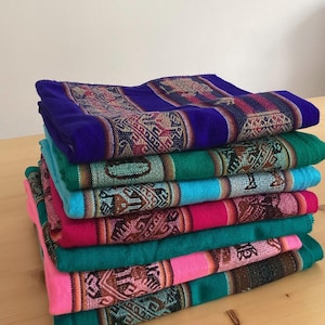 May include: A stack of colorful woven textiles. The textiles are in shades of blue, green, pink, and turquoise. Each textile has a patterned border with geometric designs in various colors. The textiles are stacked on a light-colored wooden surface.