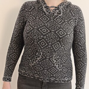 May include: A gray and black patterned hooded sweater with a front pocket. The sweater features a geometric design and a drawstring closure at the neckline. The long-sleeved sweater is made of a knitted material.