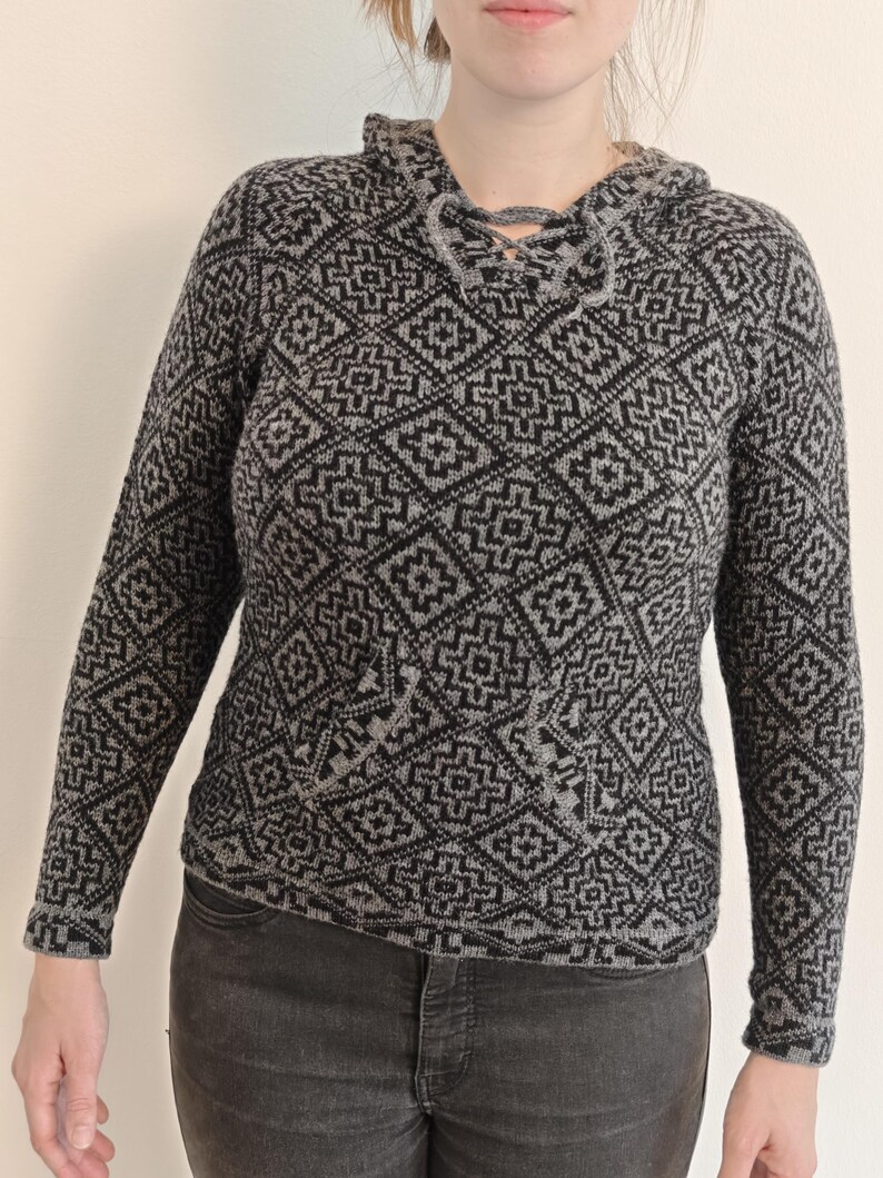 May include: A gray and black patterned hooded sweater with a front pocket. The sweater features a geometric design and a drawstring closure at the neckline. The long-sleeved sweater is made of a knitted material.
