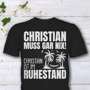 Könnte beinhalten: Schwarzes T-Shirt mit weißem Text "CHRISTIAN MUSS GAR NIX! CHRISTIAN IST IM RUHESTAND" und einer Grafik von Palmen und einer Hängematte. Das Shirt wird auf einer hellen Holzoberfläche mit weißen Blumen präsentiert.