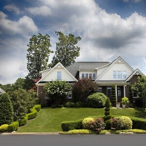 May include: A detached house with a white and brick facade, set in a landscaped garden. The house features a dark grey roof and several windows. The sky is filled with fluffy white clouds.