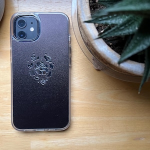 Könnte beinhalten: Eine transparente Handyhülle mit dunkelviolettem Rücken. Die Rückseite der Hülle hat ein Blumenmuster. Die Handyhülle liegt auf einer Holzoberfläche. Die Handyhülle ist für ein iPhone konzipiert.