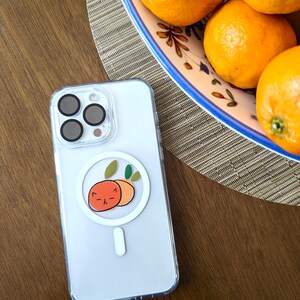 May include: A white phone case with a clear back and a cute orange sticker on the back. The sticker features two oranges with a face on one of them. The phone case is sitting on a wooden table next to a bowl of oranges.