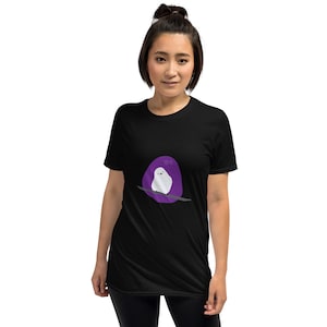 May include: A black t-shirt with a purple circle and a white bird sitting on a branch. The bird is looking to the left and the text "Hiya" is written above it.