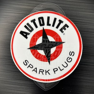 May include: Circular sticker featuring the text "AUTOLITE SPARK PLUGS" in black and white. The design includes a red and black starburst emblem at the centre, framed by a red border. Set against a dark, textured backdrop.