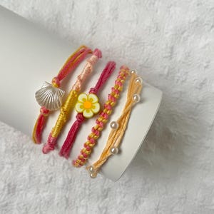 May include: A set of five colourful string bracelets featuring a white shell charm, a yellow and white flower bead, and a peach-coloured cord accented with small white faux pearls. The bracelets are crafted from pink, yellow, and peach threads.