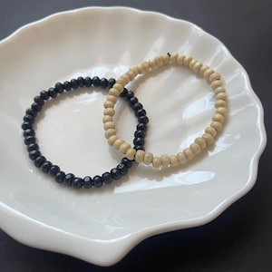 May include: Two circular beaded bracelets, one featuring small, smooth black beads and the other composed of light beige, natural-toned wooden beads. Both bracelets are constructed with a flexible, elastic cord, allowing them to maintain a round shape.