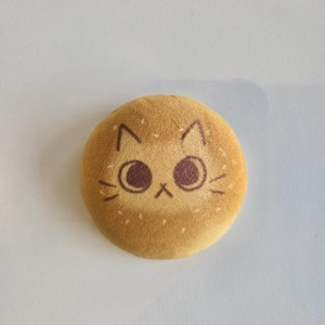May include: A round, tan-colored plush cat face with brown eyes, whiskers, and ears. The plush has a textured surface and is set against a white background. The cat's face is centered on the plush.