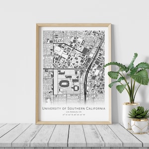 May include: A framed black and white map print of the University of Southern California in Los Angeles, CA. The print includes the university's location coordinates. The frame is a light wood color, and the print is displayed on a white surface.