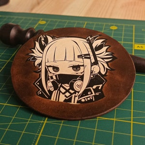 May include: A brown leather coaster featuring a white and black illustration of an anime character wearing a gas mask. The character has long white hair with black streaks and is wearing a black jacket with a white collar.
