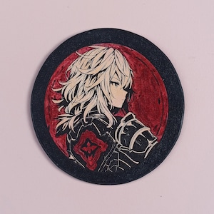 May include: Circular coaster with a side profile illustration of a character with blonde hair, set against a red circle and black border. The character is wearing black armour with a red emblem. The coaster is likely made of wood or a similar material.