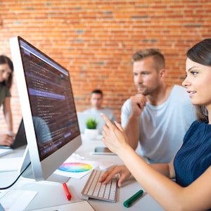 May include: A group of people working together in an office setting. A woman points at a computer screen displaying code, while others observe. A keyboard, pens, and a color wheel are on the desk. The background features a brick wall.