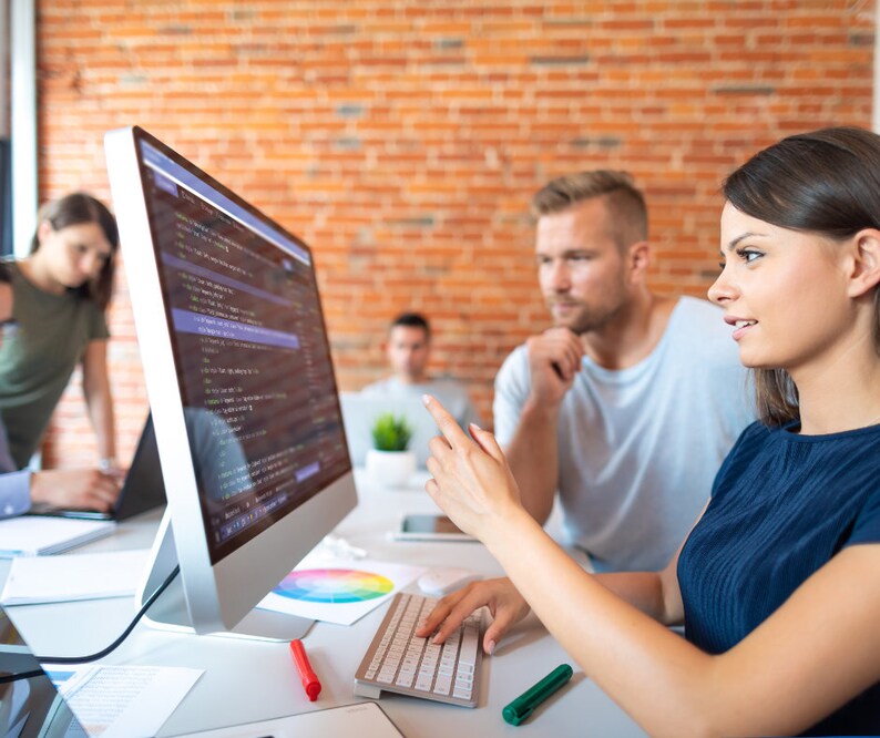 May include: A group of people working together in an office setting. A woman points at a computer screen displaying code, while others observe. A keyboard, pens, and a color wheel are on the desk. The background features a brick wall.