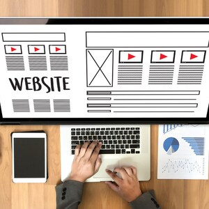 May include: Overhead shot of a laptop and tablet on a wooden desk. The laptop displays a website wireframe with the word "WEBSITE" in large text. A person's hands are typing on the laptop keyboard. A cup of coffee, glasses, and charts are also on the desk.