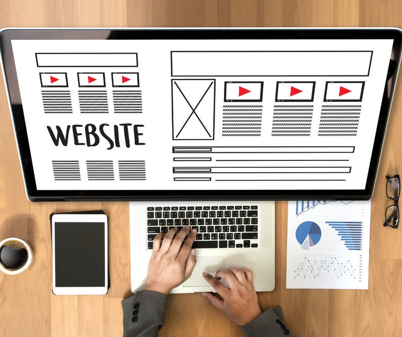 May include: Overhead shot of a laptop and tablet on a wooden desk. The laptop displays a website wireframe with the word "WEBSITE" in large text. A person's hands are typing on the laptop keyboard. A cup of coffee, glasses, and charts are also on the desk.