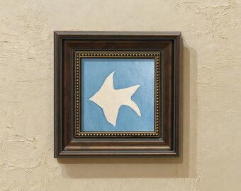 Op de afbeelding: Een vierkant wandkunstwerk met een wit abstract vissilhouet in het midden op een lichtblauwe achtergrond. Het kunstwerk is gevat in een donkerbruine houten lijst met een decoratieve binnenrand van kleine goudkleurige metalen noppen.