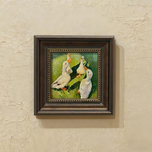 May include: A framed painting depicts four white ducks in a grassy field. The ducks have orange beaks and feet. The painting is in a dark brown frame with a gold beaded inner border. The background is a light beige wall.