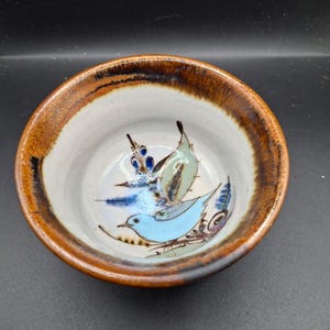 Könnte beinhalten: Kleine, runde Keramikschale mit braunem Rand und weißem Innenraum. Die Schale zeigt ein handgemaltes Design eines hellblauen Vogels mit grünen und blauen Akzenten. Die Schale wird wahrscheinlich zum Servieren von Speisen oder als dekoratives Stück verwendet.