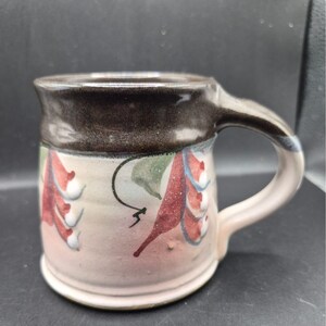 Op de afbeelding: Een handgemaakte keramische mok met een bruine rand en oor. De mok heeft een lichtroze body met rode en groene bloemmotieven. Het oor is lichtroze met een vleugje blauw. Perfect voor koffie of thee.