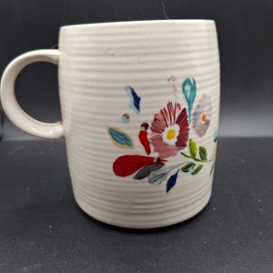 K&ouml;nnte beinhalten: Cremefarbene Keramiktasse mit floralem Design. Die Tasse hat eine gerippte Textur und einen gebogenen Griff. Das florale Design umfasst rote, rosa, blaue und gelbe Blumen mit gr&uuml;nen Bl&auml;ttern. Geeignet f&uuml;r hei&szlig;e oder kalte Getr&auml;nke.