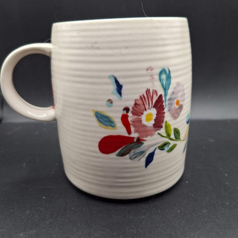 K&ouml;nnte beinhalten: Cremefarbene Keramiktasse mit floralem Design. Die Tasse hat eine gerippte Textur und einen gebogenen Griff. Das florale Design umfasst rote, rosa, blaue und gelbe Blumen mit gr&uuml;nen Bl&auml;ttern. Geeignet f&uuml;r hei&szlig;e oder kalte Getr&auml;nke.