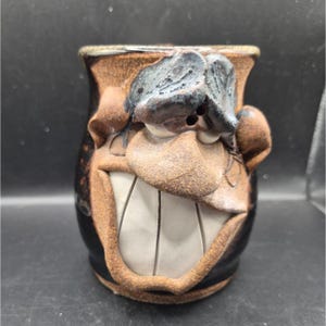 May include: A handmade ceramic mug with a whimsical face design. The mug features a smiling face with large teeth, a prominent nose, and textured details. The color palette includes brown, black, and white, creating a unique and expressive piece.