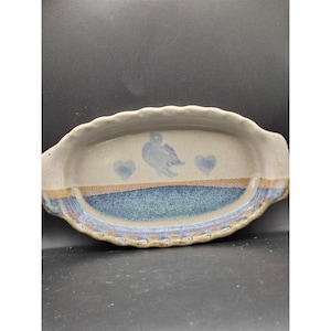 Puede incluir: Fuente de cerámica hecha a mano con borde festoneado. El plato presenta un diseño de pájaro azul y corazones sobre un fondo crema. Una banda azul y marrón separa la parte superior crema de la base azul. El plato mide aproximadamente 30 cm de largo.