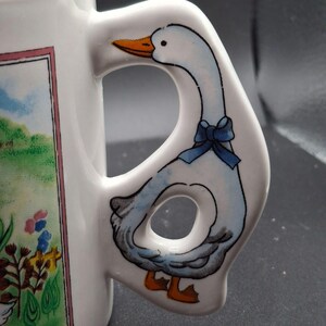 May include: White ceramic mug with a goose-shaped handle. The handle features a blue bow and a goose with orange feet and beak. The mug has a colorful illustration of a landscape with flowers and a goose.