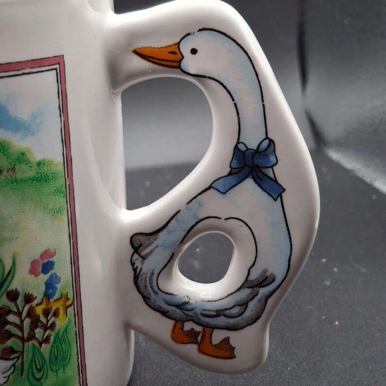 May include: White ceramic mug with a goose-shaped handle. The handle features a blue bow and a goose with orange feet and beak. The mug has a colorful illustration of a landscape with flowers and a goose.