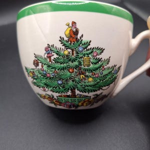 Op de afbeelding: Witte keramische theekop met een groene rand en een oor. De kop heeft een kerstboomontwerp met de kerstman, ornamenten en cadeaus. Het ontwerp is in groen, rood, geel en blauw.