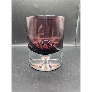 Puede incluir: Un vaso de vidrio transparente con un diseño de dos tonos. La mitad superior es de color púrpura intenso, mientras que la mitad inferior es transparente. La base presenta un diseño redondeado y bulboso. El vaso probablemente se usa para beber.