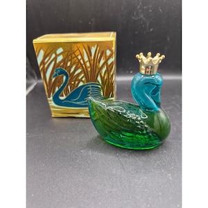 May include: A vintage glass swan perfume bottle with a gold crown stopper. The swan is teal and green, with a matching box featuring a swan illustration in teal and gold. The box has a gold background with a reed design.